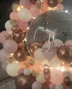 Pink white and golden balloon garland with add on white flowers wrapped around a clear balloon that has text Happy birthday love finally decorated with fairy lights